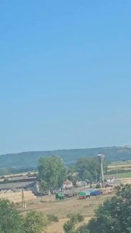 The video captures a panoramic view of a rural landscape under a clear blue sky. In the foreground, green foliage and dry grass are visible. As the camera pans, a cluster of farm buildings with red-tiled roofs comes into view, alongside stacks of hay bales. Further in the distance, a rolling green hill forms the horizon. The scene appears to be a sunny day in the countryside.