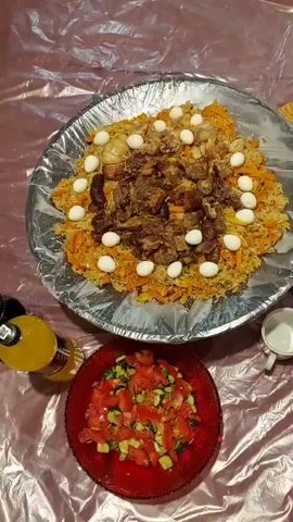 A large platter of pilaf, topped with meat and quail eggs, sits on a table covered with a crinkled, metallic-like tablecloth. Beside the platter is a red bowl filled with a tomato and cucumber salad, and a bottle of yellow liquid. The scene is filmed from above.