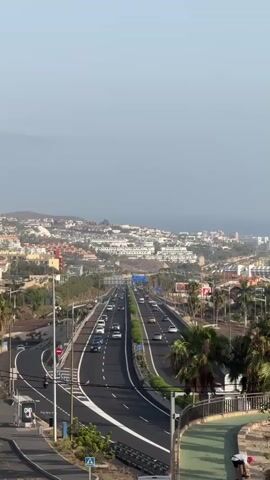 The video is an overhead shot of a highway with cars driving in both directions, a city in the background, and a walking/biking path in the foreground. There are palm trees and other vegetation along the highway and in the city. In the foreground, there are two people on the walking/biking path. One person is kneeling and appears to be looking at something on the ground. The other person is standing and appears to be looking at the same thing. The sky is clear and the lighting suggests it is daytime, possibly late afternoon or early evening due to the angle of the sun.