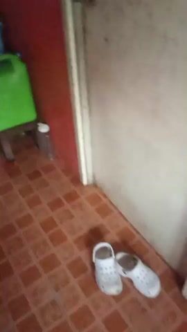 The video shows an indoor scene, likely a kitchen or dining area, with tiled flooring and red walls. Several water containers, including a large green one with a dispenser, are visible. There are also plastic bottles, a white cooler, and what appears to be a refrigerator. In the latter part of the video, a young girl with brown hair tied back is seated at a red table, looking down at something on the table. A white plastic cup is in front of her. The scene appears to be dimly lit, possibly due to natural light coming from a window.