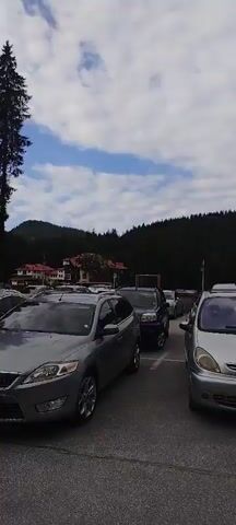 The video shows a parked cars on a cloudy day. The cars are parked in a lot in front of a forest. The forest is dense and appears to be on a hill. The sky is blue with some white clouds. The video pans to the right, showing more cars and trees.