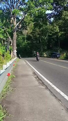 The video shows a paved road with a sidewalk beside it, flanked by trees and foliage. Two motorcycles pass by on the road, traveling in the same direction, away from the viewer. A parked car is visible on the far side of the road, near the edge of the wooded area.