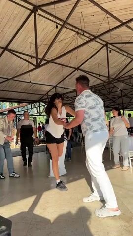 This is a video of a couple dancing under a wooden-beamed pavilion. The woman wears a white tank top and black shorts, while the man is in a white patterned shirt and white pants. They are holding hands and appear to be enjoying themselves. Other people are visible in the background, some standing and others dancing. The floor is tiled, and there are tables and chairs set up, suggesting an outdoor event or gathering. The lighting suggests it is daytime.