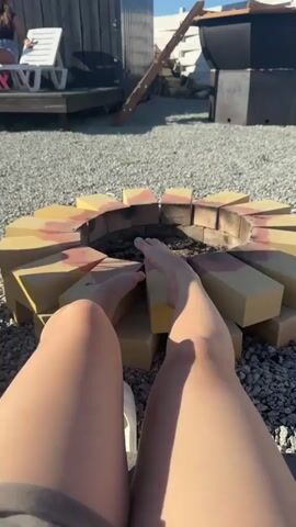 A camera shows someone's legs and feet relaxing by a brick fire pit on a gravel-covered ground. The fire pit is ringed with tan bricks. Behind the pit is a metal cauldron on a stand, with a chimney rising.  A small wooden shed is visible in the background, along with a white fence. The camera then pans up to reveal magenta-colored wooden structures in the background, set against a clear blue sky. The camera continues to pan across the gravel area to show more of the fence.