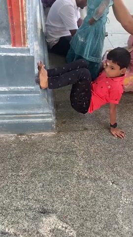 A young boy in a red shirt and black pants is doing a crab walk on a grey concrete surface next to a blue and grey stone structure. His feet are propped against the structure, and his hands are on the ground. Behind him, other people are visible, including a woman in a blue dress and a man in a white shirt. The environment appears to be outdoors, possibly near a temple or shrine, given the decorative elements on the structure and the presence of other people in traditional attire. The lighting suggests it is daytime.