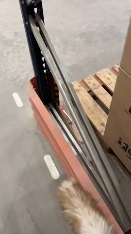 A close-up shot shows a person's arm and hand holding a dusting wand, actively sweeping it across a metal rack system. The wand is blurred due to motion, indicating vigorous movement. The rack appears to be part of a storage or shelving unit, with orange-colored supports visible at the bottom. To the right of the rack, a wooden pallet with a cardboard box on it is partially visible. The flooring is a gray, concrete-like surface, with two yellow markings. The overall scene suggests a cleaning or maintenance activity in an industrial or warehouse setting.