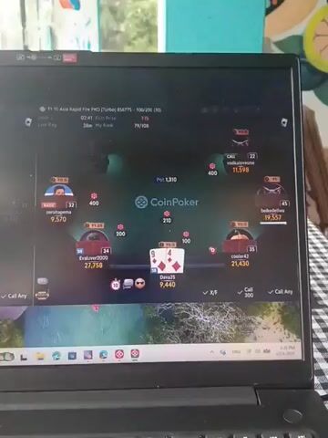 The media file is a close-up, eye-level shot of a laptop screen displaying an online poker game. The screen is divided into two main sections, showing two separate poker tables.

On the left table, the pot is 7,888. The player "Romantics" has 6,807 chips, and their cards are an 8 and a 7 of hearts. The player "Dava25" has 2,491 chips. The community cards are a King of spades, a 9 of hearts, and a 2 of spades. The time stamp at the top indicates 01:12 and "My Rank" is 48/96.

On the right table, the pot is 1,710. The player "zerotapema" has 9,570 chips, and their cards are a 10 of spades and a 5 of spades. The player "Evaluvc2000" has 27,758 chips. The community cards are a 9 of hearts, a 4 of spades, and a 4 of diamonds. The player "Dava25" has 9,440 chips. The time stamp at the top indicates 02:39 and "My Rank" is 79/108. The table is labeled "T1.10 Asia Rapid Fire PK4 (Turbo)".

The laptop is open and appears to be on a table or surface. In the background, out of focus, there are glimpses of colorful walls or artwork, suggesting an indoor setting. The image quality is slightly grainy. The lower part of the laptop shows a taskbar with various icons and a search bar, with the date and time "04/14/2020 9:26 PM" visible in the bottom right corner. The overall scene depicts someone engaged in playing online poker. The location context is Dharamshala, India, though this is not directly visible in the media, it is implied by the prompt.