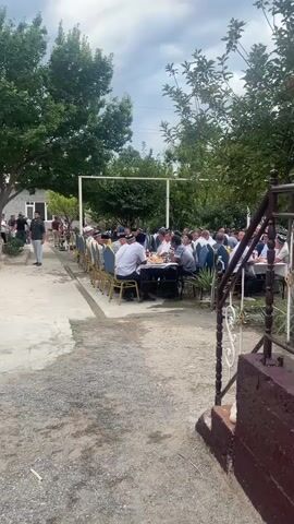 The video shows an outdoor gathering with many people seated at long tables under trees. The tables are laden with food and drink. Some people are standing and conversing in the background, while others are walking along a path. The environment appears to be a courtyard or garden, with buildings and vegetation visible. The weather seems to be overcast.