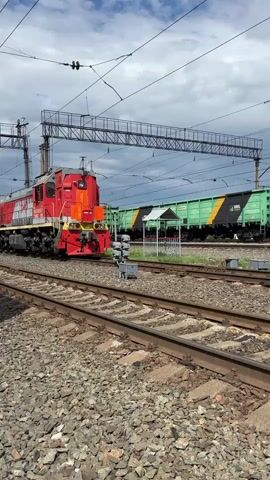 A red and white locomotive is stationary on a railway track, positioned slightly to the left of the frame. It has the letters "РЖД" in red on its side. Behind the locomotive, several green freight cars are visible on another track, with some of them partially obscured by the locomotive and other structures. The ground is covered with ballast stones. Overhead, power lines and supporting structures are visible against a cloudy sky. The camera is focused on the train and tracks, with the background slightly blurred.