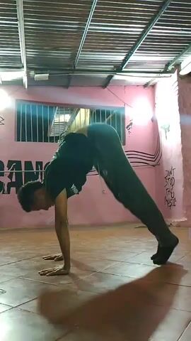 A male individual performs a breakdancing move in an indoor setting, likely a dance studio or practice room in El Espinal, Venezuela. He is wearing a black t-shirt and dark pants. The room features pink-painted walls with graffiti-style art, including the large word "DANCE" and the phrase "DON'T STOP THE MUSIC." The floor is tiled, and the ceiling has an industrial-style metal structure. Artificial lighting illuminates the space. The individual initiates a movement from a low stance, transitions into a handstand, and then executes a controlled fall onto his back, ending in a semi-bridge pose with his hands covering his face, indicating a complex acrobatic maneuver. Music is playing in the background.