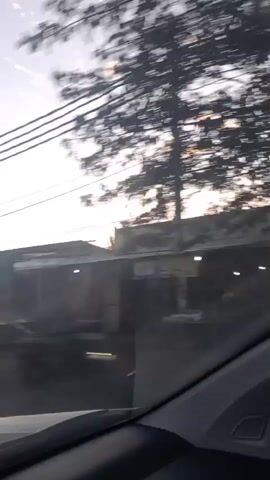 The video captures a blurry, moving perspective of an outdoor environment during what appears to be dawn or dusk. Power lines are prominently featured crisscrossing the frame. In the background, dark, indistinct buildings and structures are visible. Trees with dark foliage are also present, contributing to the overall shadowed atmosphere. The sky is a pale blue with scattered clouds, some of which are illuminated, suggesting the sun is low on the horizon. The view suggests the footage is recorded from a vehicle moving along a street or road.