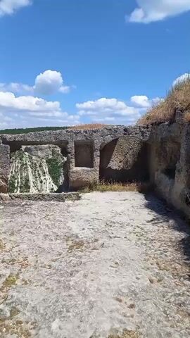 The video pans across an ancient stone structure under a bright blue sky with scattered white clouds. The structure appears to be carved out of rock, with rough-hewn walls and openings. In the center, a large rectangular opening reveals a view of a rocky landscape with some greenery, possibly a canyon or a steep hillside, with what looks like a waterfall effect overlaid on it. To the left of this opening, there's a rough, stepped structure also carved into the stone. The foreground is a cracked, stony ground with sparse dry grass. To the right, another rectangular opening is visible. The camera moves to the left, revealing more of the stone structure's exterior, including more rough surfaces and openings. The overall impression is of an ancient ruin in a natural, open environment.