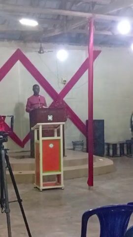 Here is a description of the video.
The video appears to be shot inside a church. A man in a pink shirt is standing at a podium, seemingly speaking. Behind him, the wall is decorated with a large red geometric design, and a monitor is visible at the top. There are other people in the room, including a woman wearing a bright green vest and other individuals in black clothing. Several items such as a tripod with a camera, fans, and speakers are visible. The scene has a slightly makeshift feel, and it looks like a service is being recorded. There is music playing in the background.