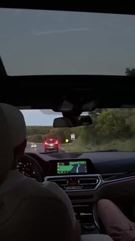 The video captures the interior of a car from a passenger's perspective, looking forward. A driver wearing a light-colored baseball cap is visible from behind, gripping the steering wheel. The car's dashboard displays a navigation system with a route highlighted in green. Ambient interior lighting illuminates the console with a purple glow. Outside the car, a road winds through a tree-lined landscape. Several cars are visible on the road ahead, their taillights illuminated. The sky appears overcast, suggesting it might be dusk or a cloudy day. The video appears to be a continuous shot, showing the car in motion.