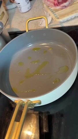 A person uses tongs to put a large, raw, red steak into a light blue frying pan with golden handles on a black stovetop. There are a few small puddles of oil in the pan. There is a container of bacon in the background, as well as a white container with a black lid.