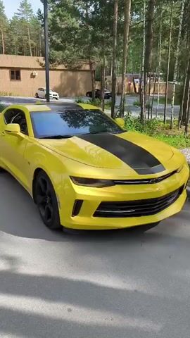 The video shows a yellow Chevrolet Camaro with black racing stripes parked on a paved surface. The car is positioned at an angle, with its front facing slightly towards the right of the frame. It is outdoors, with a backdrop of tall trees and a building visible in the distance. The lighting suggests it is daytime, possibly late afternoon due to the shadows. There are no people visible in the immediate vicinity of the car, and no actions are taking place. The environment appears to be a natural or semi-natural setting, possibly a private driveway or a park area.