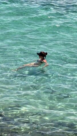A woman floats on her back in crystal clear, aquamarine water, wearing a black swimsuit and a dark baseball cap. Her hair is tied up in a bun. Ripples spread across the water's surface, and sunlight reflects off the seabed below, creating patterns of light and shadow.