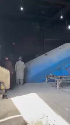The video shows the inside of a dark and dusty warehouse. In the background is a large, blue, cylindrical tank tilted at an angle. Two men stand to the left of the tank. One man wears a brown robe and the other a light gray robe. The floor is made of wooden planks. Overhead are bare light bulbs. The walls are brick, and appear dirty or burnt. The tank is resting on wooden supports and sand. There are also metal pipes in the foreground. The video seems to be taken from a handheld device. Sounds of machinery can be heard.