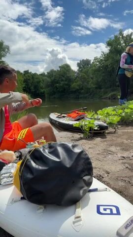 Three people are outdoors next to a body of water under a partly cloudy sky. In the foreground, part of a white inflatable paddleboard with a black dry bag rests on a sandy bank. To the left, a person wearing an orange vest is seated, partially visible, and appears to be eating from a metal container with a spoon. On the right, two women are standing on the sandy bank near a patch of green leafy plants. The woman on the left, wearing a patterned long-sleeved top, black leggings, a yellow fanny pack, and a blue visor, bends down to interact with the plants or ground, then stands up holding a small item. The woman on the right, in a pink long-sleeved top and a pink baseball cap, also bends down and then stands, bringing her hand to her mouth. In the mid-ground, a black inflatable paddleboard with an orange life vest on it rests partially in the water beside the bank. The background consists of a calm river and a dense line of green trees.