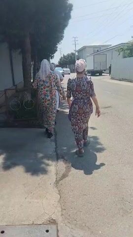 Two women wearing matching colorful patterned dresses and head coverings walk along a sidewalk next to a street. One woman is wearing a white headscarf that covers her hair and part of her face.  A dark green car passes by on the street.  There is a fence next to the sidewalk, trees, and buildings in the background.  The weather is sunny.