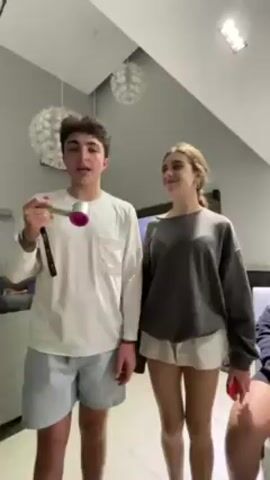 A boy and a girl are standing side by side indoors. The boy is holding a pink and silver hairdryer up to his mouth and pretending to sing into it. Both are smiling and opening their mouths as if singing. The boy is wearing a white long-sleeve shirt and blue striped shorts. The girl is wearing a gray sweatshirt and white shorts, holding a phone in her right hand.  In the background, there is a gray wall with two hanging light fixtures and a dark entertainment center to the right. The lighting is bright.