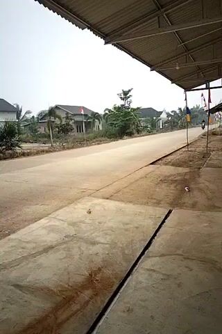 A person is riding a motorcycle down a road. The motorcycle is black and the person is wearing black clothing and a helmet. The road is paved and appears to be in a rural area. There are houses and trees on either side of the road. The sky is overcast. The video is taken from a low angle, looking up at the road and the motorcycle.