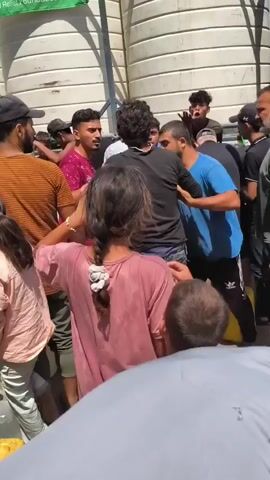 A group of people are gathered outdoors, some engaged in a physical altercation. One man in a blue shirt is arguing with another man in a black shirt. Several other individuals are attempting to separate them and de-escalate the situation. There are children present, and some people are holding containers. The background shows a large white cylindrical structure and a green banner with text. The ground appears to be dirt.