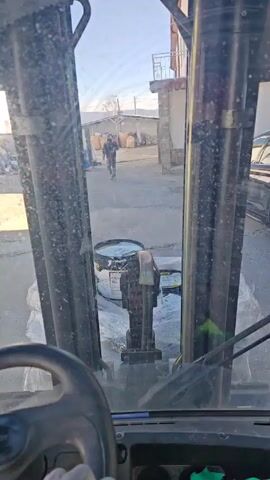 The video shows a view from inside a forklift moving slowly forward in what appears to be an industrial or storage yard.  A large black drum and some bags covered in clear plastic are on the forklift. The driver, wearing black gloves and a red jacket, is operating the vehicle. Through the dirty windshield, a person in dark clothing is walking away in the distance. Other buildings and stacked items are visible in the background under a bright sky. A black SUV is partially visible on the right side. The camera is fixed and stable, capturing the driver’s perspective.
