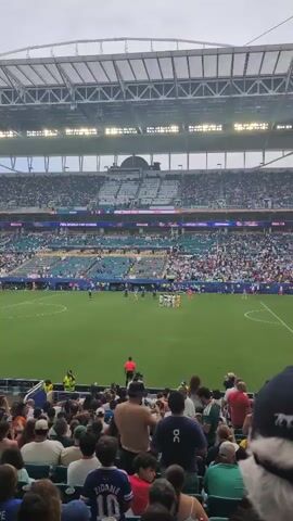 This video shows a soccer match in a stadium with a large crowd of spectators. The field is green grass, and there are players on both sides of the field, one team in white uniforms and the other in blue uniforms. The spectators are seated in tiered rows and are looking towards the field. The stadium has a large, white, domed roof. The weather appears to be cloudy. The video is shaky, suggesting it was recorded handheld.
