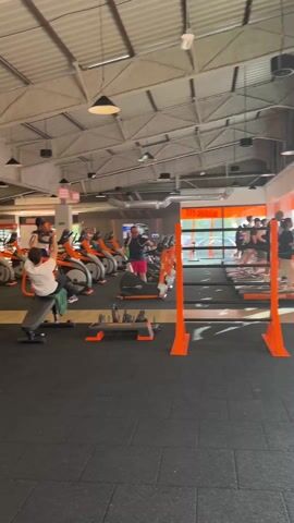 The video shows the interior of a well-lit gym with black rubber flooring and a high, angular ceiling with visible metal support beams. Multiple individuals are present, engaging in various exercises on different types of equipment.  There are rows of stationary bikes and elliptical machines, most colored orange and black. People are using these machines. A person is using a weight bench. Another person is near the floor.  The overall atmosphere is one of active fitness engagement.