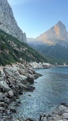 The video begins with a panoramic view of a rocky coastline with a calm blue sea. Steep, rocky hills covered in sparse greenery rise dramatically from the water's edge. In the distance, a prominent, sun-kissed mountain peak dominates the horizon. The camera then pans across the water, revealing more of the rugged coastline. As the view widens, two individuals can be seen standing on the rocks by the sea, facing the water. The camera continues to zoom out, showing the vast expanse of the ocean meeting the clear blue sky. The last shot shows a wider view of the coastline with the ocean stretching out to the horizon.