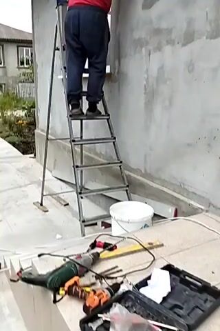 A person wearing a red shirt, blue pants, and a white cap stands on a metal ladder against a gray concrete wall. The person is using a tool to work on the wall, possibly plastering or repairing it. The ladder has several steps, and the person is on the fourth step from the bottom. There is a window to the left of the ladder. In the foreground, there are tools and a white bucket. The environment appears to be an outdoor construction site.