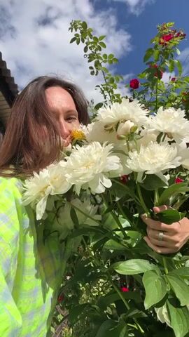 A woman with long brown hair holds a large bouquet of white peonies and sniffs them. She is wearing a green and yellow checkered shirt. Behind her, a house with beige siding and a rose bush with red roses can be seen. The sky is visible above, with white clouds. The woman's face is partially obscured by the flowers, but she appears to be smiling. She is holding the bouquet with both hands, and her left hand is resting on the stems. A ring is visible on her left ring finger. The overall scene suggests a warm, sunny day in a garden setting.
