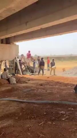 The scene unfolds beneath a concrete bridge. Several individuals are engaged in construction work. One person, wearing pink, stands atop a concrete mixer while others attend to it. Other workers, some in hard hats, are scattered around a pile of gravel, carrying buckets. The ground is a mixture of red soil and scattered debris. A blue pipe snakes across the foreground. The background is a hazy landscape of dry vegetation.