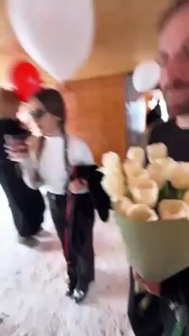 The video shows a group of people indoors. A woman wearing sunglasses, a white long-sleeved shirt, and braided hair takes a sip from a paper cup. She is holding a red balloon. A man to her right holds a bouquet of white roses. Other people are standing in the background. The environment appears to be a building or covered area with wooden beams. A white balloon floats in the air. The floor is light-colored.