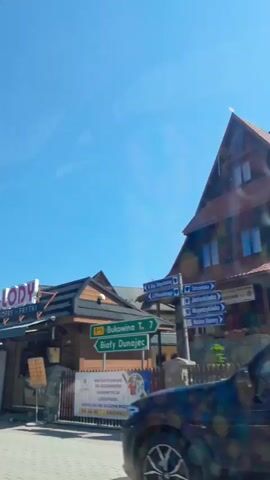 The video begins with a street view of several food stalls with awnings and signs that read "LODY" which translates to "ice cream" in Polish. People are seen standing in line at the stalls. The camera then pans to the right, showing a street with a clock tower, a church, and several traditional wooden buildings with steep roofs. A car is seen driving down the street. The view shifts again to show a large evergreen tree with a bench underneath it, as well as a white car parked on the side of the road. The sky is clear and blue throughout the video.