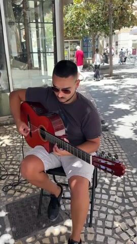 A young man with dark hair, a beard, and sunglasses is playing a red acoustic guitar while sitting on a black folding chair outdoors. He is wearing a dark t-shirt and white shorts. A small black amplifier with red accents is positioned to his left. A guitar case rests on the ground next to the amplifier. The man is seated on a tiled walkway in front of a storefront with large glass windows. Several people are walking in the background on the street, and trees provide shade. The man is actively strumming and fretting the guitar, indicating he is performing.