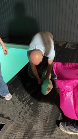 The video shows a man in a light blue shirt and jeans bending over to hold a child's inflatable pool toy. Next to him on the deck is a deflated pink inflatable lounge. Another man with a dark beard holds the deflated lounge at his side. A woman standing near the side of a glowing blue pool has her hand raised in a gesture, but is not speaking. The men appear to be trying to assemble the pool toys. The setting appears to be an outdoor patio at night.