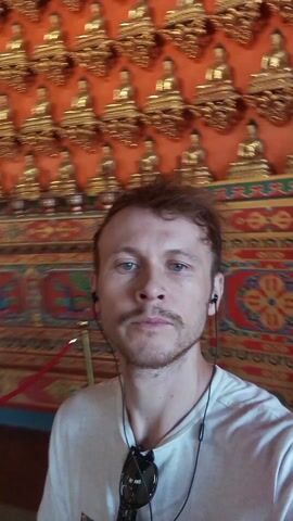 The video features a man in his late 20s to early 30s with light brown hair, a mustache, and a slight stubble beard. He is wearing a white T-shirt and has earphones in his ears. Sunglasses hang around his neck. He appears to be recording a video of himself.

Behind him is a wall decorated with numerous small, golden Buddha statues arranged in a grid pattern. The wall's background is a warm orange color. Below the Buddha statues is a more elaborately decorated panel with intricate designs featuring figures and animals, predominantly in blue, red, and gold colors.

The man is looking directly at the camera, speaking, though the audio is not transcribed here. The camera remains relatively static, focusing on the man and the background. The lighting is fairly bright, suggesting the scene is well-lit. The overall impression is that the man is inside a temple or a religious building with significant Buddhist ornamentation.