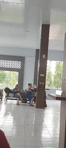 The video shows an indoor waiting area with tiled floors and large windows. Two women are seated on a bench, both looking at their phones. A third person, a man, is standing and looking towards the left. There are bags on the floor and on the bench. The overall lighting is bright and natural.