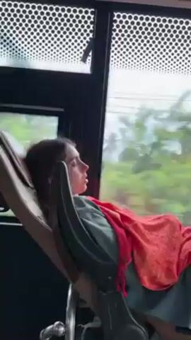 A person, likely a young woman, reclines on a special chair aboard a moving vehicle, possibly a bus, as suggested by the window with small, round perforations above and the passing scenery featuring green trees. The individual is lying back with their eyes closed, a red shawl draped over them.  The specialized chair appears to be adjustable, with levers and a headrest.  The overall lighting is diffused, and the scene seems captured from a slightly upward angle.