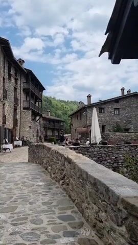 The video shows a quaint European village street paved with cobblestones and lined with stone buildings. On the left, a multi-story building with dark balconies and windows faces a courtyard with white-clothed tables and chairs. A few people are seated at these tables. In the center, a stone wall separates the cobblestone path from an area with more stone buildings. One building has a white umbrella and tables with people seated outside. The background reveals a lush green hillside with trees under a partly cloudy sky. The perspective is from a low angle, looking up the street towards the hillside.