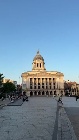 The video shows a large building in a city square. The building is pale in color and has a domed roof with a clock on it. There are columns across the front of the building, and a flag is flying from the dome. In front of the building is a large paved square with people walking around. Some people are sitting on a low wall. The sky is blue. The video zooms in on the clock on the dome of the building. The clock face is white with black numbers. The time is approximately 8:14.