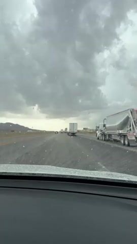 The video shows a view from inside a vehicle driving on a highway. The sky is overcast with heavy, gray clouds. The road appears to be wet, and there are scattered white specks on the surface, suggesting precipitation such as rain or hail. Several semi-trucks are visible on the highway, traveling in the same direction as the camera. The surrounding environment appears to be a dry, arid landscape with low hills or mountains in the distance. The overall mood is somber due to the weather conditions.