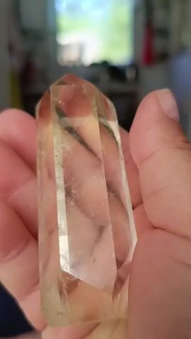 A hand holds a clear, faceted crystal with a slight yellow tint. The crystal is oriented vertically, with its pointed end facing upwards. The hand is positioned to the right of the crystal, with fingers supporting its lower portion and thumb wrapping around the side. The background is out of focus, showing a blurred interior scene with a window and some furniture. The video captures the crystal from various angles as it is rotated and tilted by the hand.