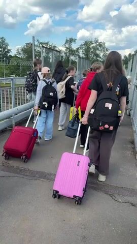 The video captures a group of young people, predominantly girls and young women, walking across an elevated pedestrian bridge at a train station in Malaya Vishera, Russia. They are pulling various colored wheeled suitcases, including red, pink, yellow, and maroon, and carrying backpacks and duffel bags.

The group appears to be in transit, possibly embarking on or returning from a journey. They walk along the concrete bridge, which overlooks multiple train tracks and railway platforms. In the background, a red and white train is visible on the tracks, along with overhead power lines and distant buildings.

The weather is bright and sunny, with a partly cloudy sky, indicating it is daytime. The mood among the group seems cheerful and engaged, with individuals talking, smiling, and occasionally glancing at the trains below. One young girl in a black hoodie with a skeleton pattern is seen smiling while looking out from the bridge. The individuals are dressed casually for travel. The bridge features metal railings on both sides, offering clear views of the railway infrastructure.