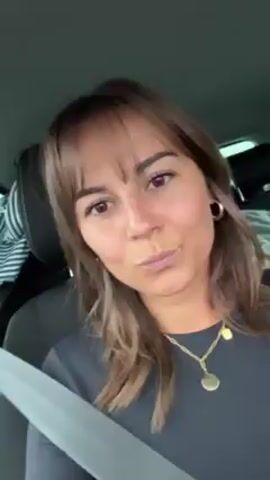 The video shows a view from the front passenger seat of a car. A woman with brown hair is filming herself. In the back seat, a woman with blonde hair appears to be asleep, leaning her head back. She is wearing glasses. A maroon-colored article of clothing appears behind her. The car is in motion.