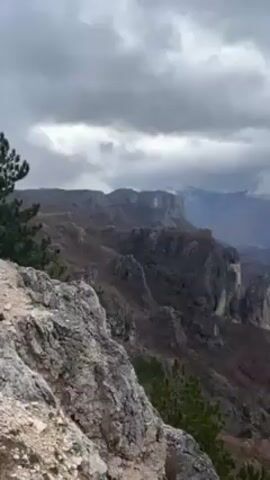 The video shows a landscape of mountains under a cloudy sky. The foreground consists of rocks and some greenery. In the middle ground, there are layers of mountains with cliffs and bare trees. Some distant mountains are visible with snow on top. The sky is overcast with heavy, gray clouds. The camera pans slightly to show more of the mountainous terrain.