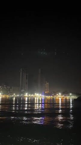 A nighttime view of a city skyline across a body of water. Tall buildings with illuminated windows are visible, reflecting in the dark water which appears to have small waves. In the distance, a faint green light pattern is present in the sky. The overall atmosphere is dark and the lighting suggests urban activity.