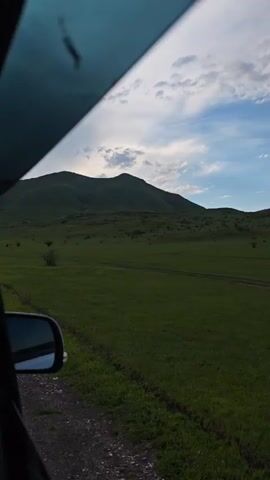 The video shows a view from the side of a car, focusing on the passing landscape. The car is traveling on a dirt road adjacent to a large, green field. The field stretches towards a distant, gently sloping mountain range, covered in lush greenery. Above, the sky is partially covered with clouds, with patches of blue visible. The overall lighting suggests late afternoon or early evening. The car’s side mirror is visible, providing a frame to the broader landscape. The scene is static, implying a simple, consistent movement.